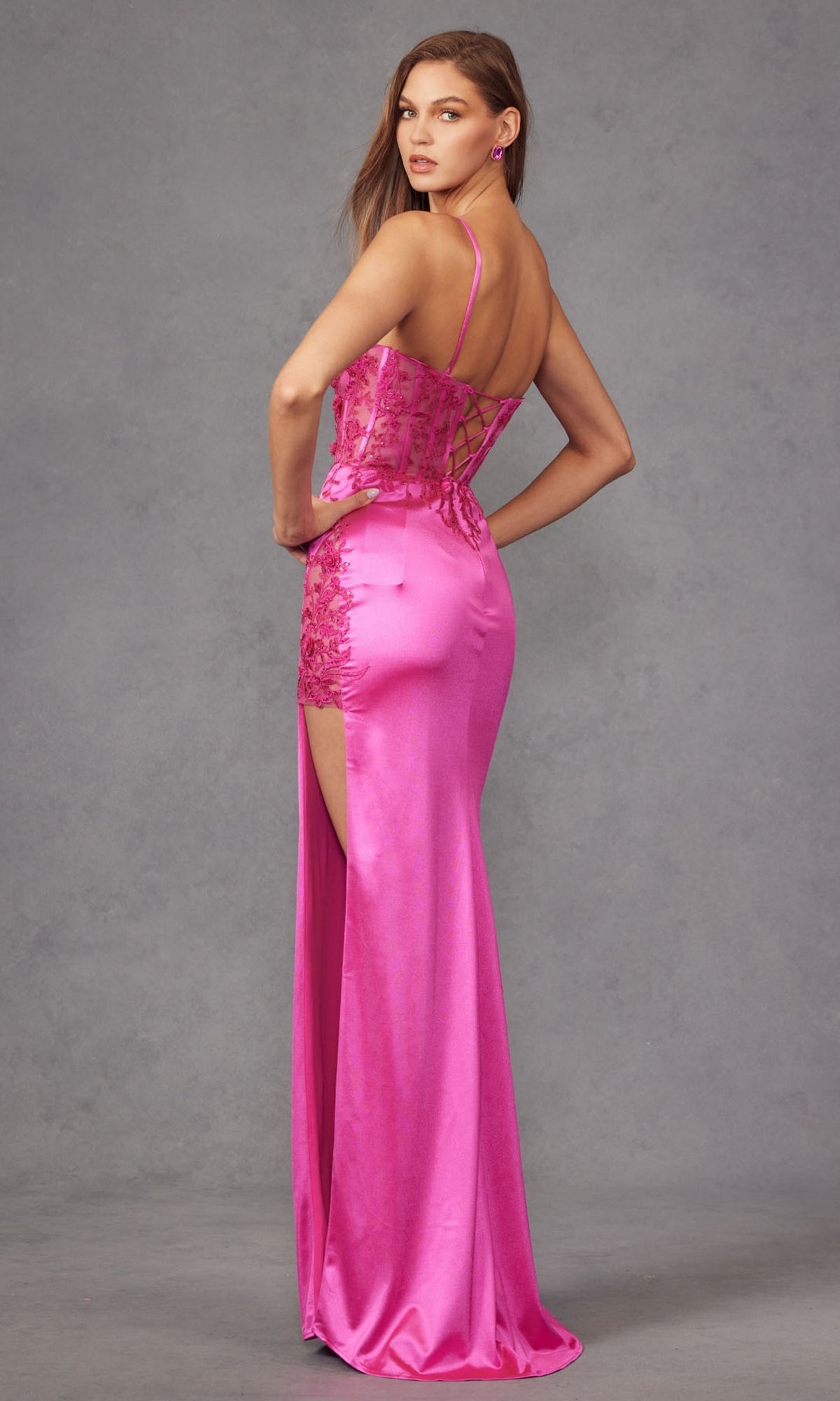 A woman models the Long Prom Dress JT2476U by Juliet—a fitted, bright pink satin gown with lace embroidery, thin straps, and a high slit—against a plain gray background.
